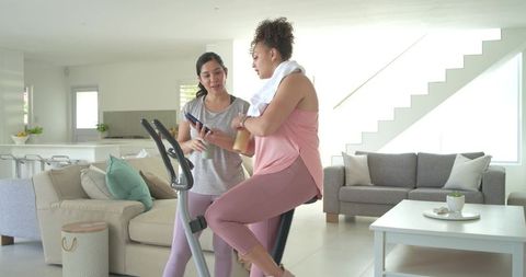Diverse women pedaling on exercise bike in bright minimalist living room checking phone