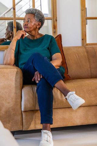 Senior african woman sitting on tan leather sofa looking thoughtful in cozy living room