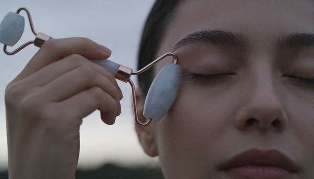 Woman rolling stone facial roller across eye at dusk for calming skincare ritual