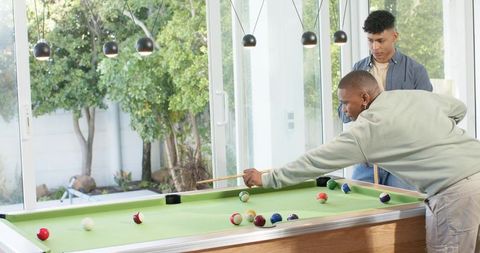 Diverse male friends playing pool in sunlit game room with glass doors and garden view