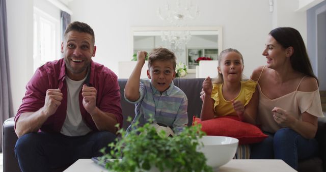 Happy family of four sitting on a sofa in a cozy living room, cheering and showing excitement while watching TV together. Ideal for commercials, advertisements, or articles about family togetherness, leisure activities, or home environments.