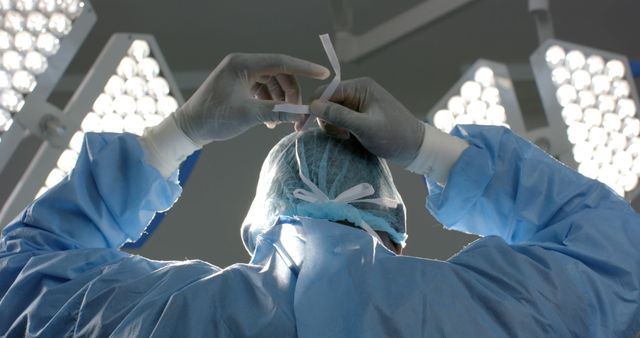 This image shows a surgeon tightening their mask under bright surgical lights in an operating room. It is ideal for use in health and medical-themed articles, websites, and promotional materials related to surgery, hospital environments, healthcare professionals, and patient care safety.