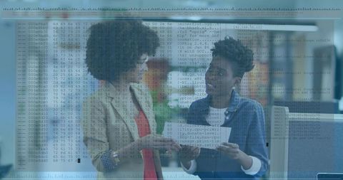 Businesswomen collaborating and reviewing documents with code overlay in modern workspace