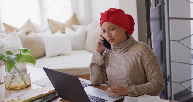 Image of smiling biracial woman in hijab at desk working on laptop talking on smartphone at home. Remote working, communication, inclusivity and domestic life.
