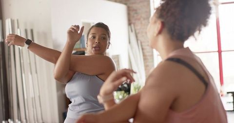 Women stretching arms across chest in mirrored industrial fitness studio with poles