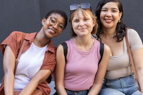 Smiling diverse female friends posing on urban wall showcasing casual summer street style