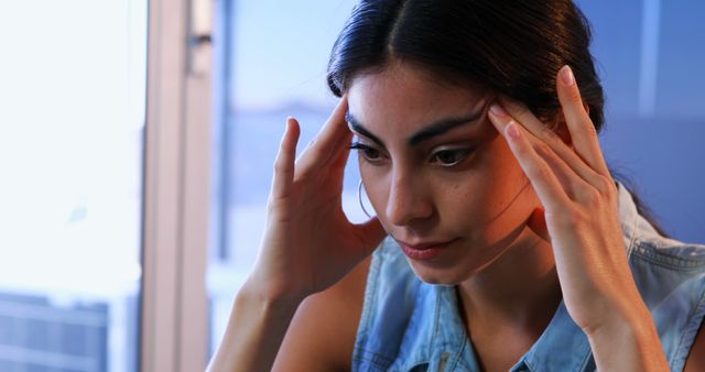 A young Hispanic woman appears stressed or deep in thought, with her hands on her temples, indicating a headache or anxiety. Her expression and posture convey a sense of concern or contemplation, highlighting the human experience of stress or worry.
