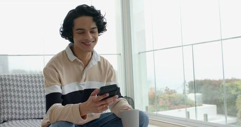 Casual young man checking smartphone on sofa with mug in minimalist bright living room