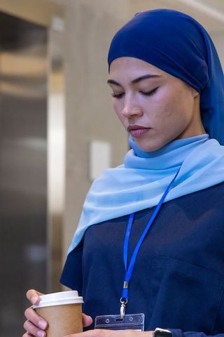 African american nurse wearing blue hijab holding coffee and id badge, contemplative portrait