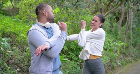 Fitness partners stretching on wooded trail wearing activewear and headphones, warming up before run