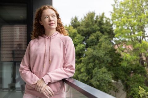 Contemplative young woman standing on balcony wearing pink hoodie, gazing outdoors