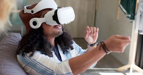 Young man wearing VR headset on sofa reaching virtual controls for immersive home gaming