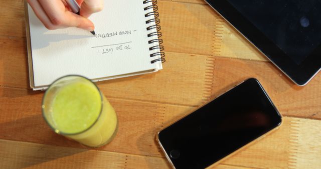 A person is jotting down notes in a notebook beside a green smoothie, with a tablet and smartphone also on the wooden table, with copy space. Capturing a moment of productivity, the scene suggests a healthy lifestyle combined with technology and work or study.
