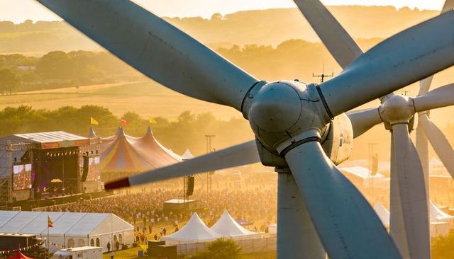 Close-up wind turbine rotor framing sunset music festival over golden farmland crowd