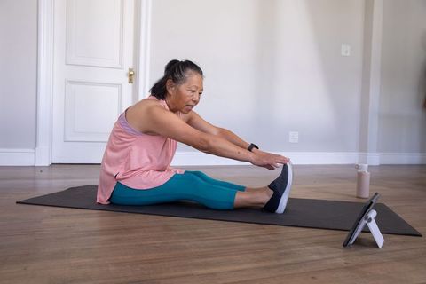 Senior asian woman stretching on mat following tablet workout in bright home interior