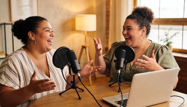 Two women laughing during cozy home podcast recording with microphones and laptop