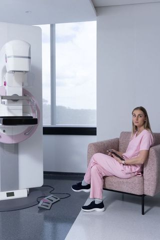 Female mammography technologist sitting in pink scrubs holding tablet in exam room