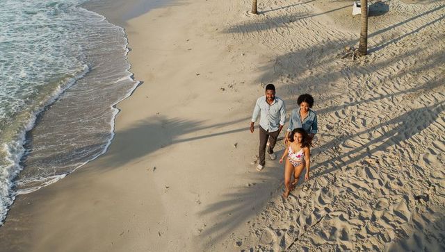 Walking family along sunlit tropical beach with palm shadows, footprints and ocean waves