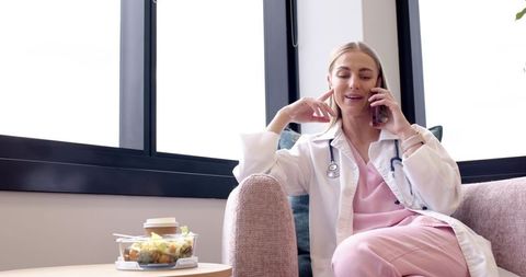 Female clinician in pink scrubs taking phone call while relaxing in modern clinic lounge