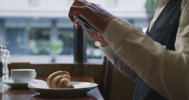 Person photographing croissant on plate with smartphone in modern cafe. Ideal for food photography, blogging, social media content, or marketing for cafes and bakeries.