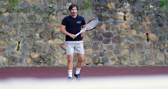 Man practicing tennis near a stone wall outdoors. He is wearing athletic clothing, including a T-shirt and shorts, and holding a tennis racket. This scene suits projects and articles related to sports training, fitness routines, athletic lifestyles, and outdoor activities.