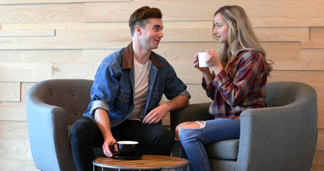 Young couple sitting in a cozy café and chatting while holding coffee cups. Man in denim jacket and woman in plaid shirt appear engaged and comfortable. Useful for depicting themes of relaxation, casual conversation, social interaction, and everyday moments. Ideal for marketing materials related to cafes, coffee brands, or lifestyle blogs.
