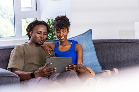 African american couple sitting on sofa sharing tablet and headphones, cozy home scene