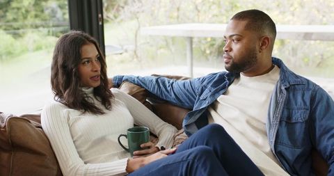 Diverse couple relaxing on leather sofa sharing intimate conversation and coffee by window