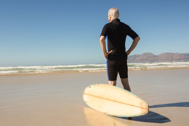 Rear view of man standing by surfboard against clear sky at beach