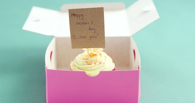 A close-up view of a cupcake inside a pink box with a handwritten message card that reads 'Happy Mother's Day, I love you!'. The cupcake features creamy frosting and colorful sprinkles, making it an ideal gift for a special occasion. Perfect for use in Mother's Day promotions, greeting cards, or advertising campaigns that involve baked goods and celebrations.