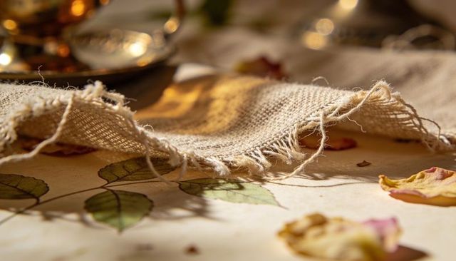 Frayed burlap cloth draping over botanical tabletop with dried rose petals and warm glassware