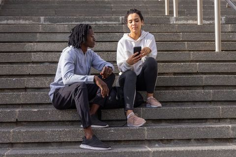 Black man and mixed-race woman sitting on concrete steps sharing smartphone and chatting casually