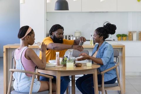 African family sharing casual kitchen meal, father seasoning salad at round dining table