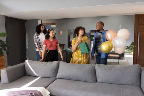 Diverse friends entering living room holding gift bag and balloons