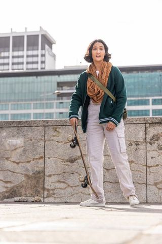 Young woman standing with skateboard on urban plaza wearing green jacket and scarf