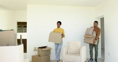 Young couple moving into minimalist living room carrying boxes and wrapped furniture