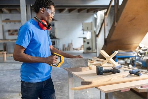 Young carpenter sawing wooden plank on workbench wearing safety goggles and ear protection