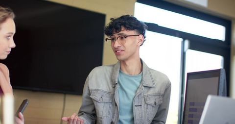 Young professional presenting in collaborative workspace wearing denim jacket and glasses