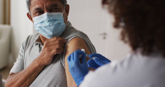 Senior man receives vaccine injection from healthcare worker in clinical environment. Senior wearing mask, healthcare professional wearing protective gloves and administering injection in arm, ensures safe medical procedure. Ideal for use in articles about vaccination, healthcare, medical services, senior care, health and wellness campaigns, and public health initiatives.