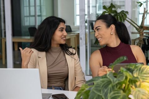 Two female coworkers collaborating at modern glass office desk with laptop and green plants
