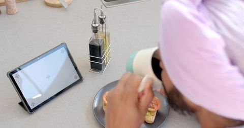Indian man in pink turban sipping coffee at kitchen counter with tablet and tomato toast