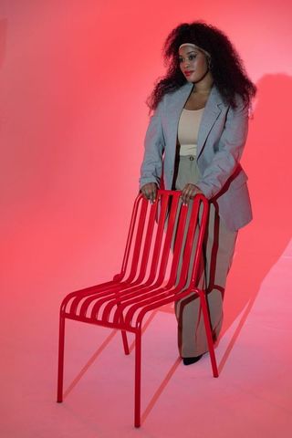 African american woman standing behind red metal chair wearing gray blazer in pink studio