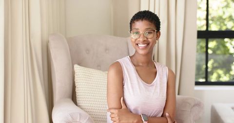 Smiling african woman wearing eyeglasses and smartwatch on beige armchair in neutral living room