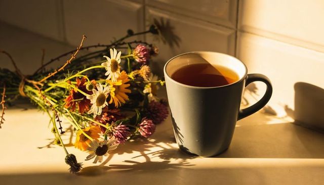 Sunlit gray ceramic mug of amber tea and wildflower bouquet casting shadows on countertop