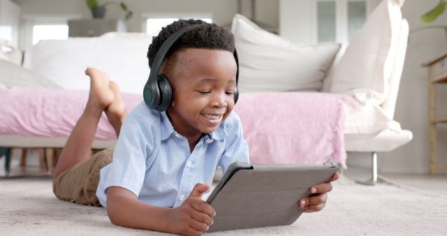 Young boy lying on floor, using tablet with headphones. Ideal for concepts like childhood, technology use, leisure activities, digital learning, listening to music or stories.