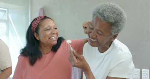 Senior african american women laughing and sharing yogurt by bathroom mirror
