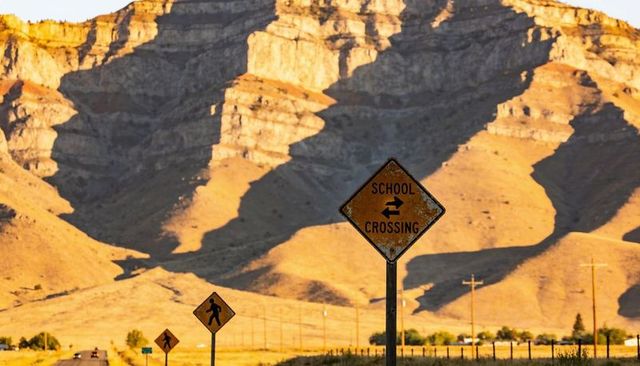 Worn school crossing sign pointing along rural two-lane road against sunlit sandstone cliffs