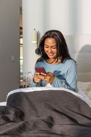 Asian woman sitting in bed smiling and texting on red phone under gray blanket in sunlit bedroom