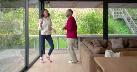 Couple dancing in bright modern living room with floor-to-ceiling glass and green view