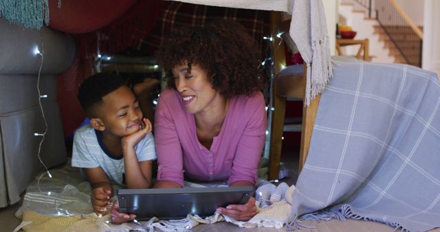 This image captures a mother and her young son enjoying time together in a homemade blanket fort, illuminated with fairy lights. Both are smiling and engaging with a tablet, suggesting screen time or entertainment. Ideal for illustrating family bonding activities, technology use in family settings, and creating playful, cozy home environments.